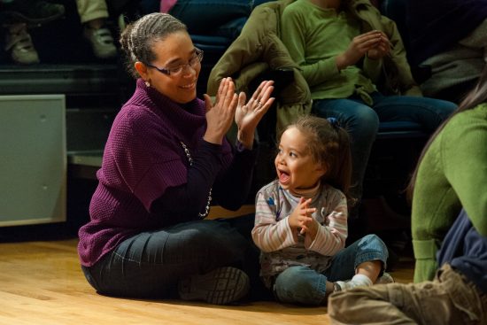 A mother sits in an audience with her young child clapping along to the music.