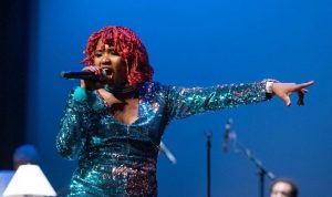 Thabisa stands with her arm outstretched, singing into a microphone. Her hair is red and she is wearing a vibrant aquamarine dress.