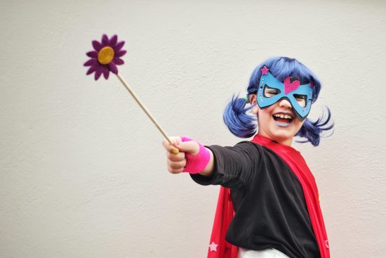 Young girl wearing a mask and super hero cape extends her right arm holding a purple gerber daisy