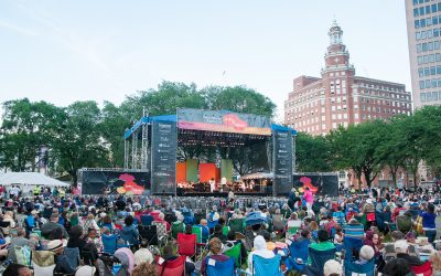 The NHSO playing onstage at the International Festival of Arts and Ideas on the New Haven green. A large crowd is visible in the foreground and the skyline of New Haven sets a backdrop behind the stage.