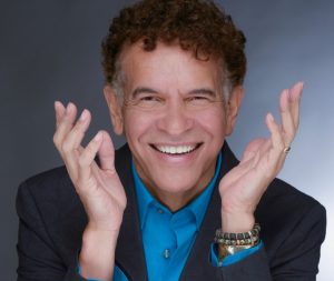 A close-up headshot of Brian Stokes Mitchell with his hands thrown playfully into the air. He smiling and looking directly into the camera.