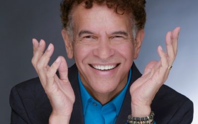 A close-up headshot of Brian Stokes Mitchell with his hands thrown playfully into the air. He smiling and looking directly into the camera.