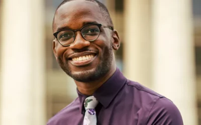Joel Thompson faces the camera smiling. He is a Black man and is wearing glasses and a purple collared shirt with a white patterned tie.