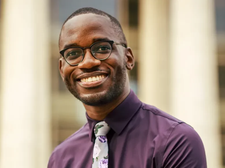 Joel Thompson faces the camera smiling. He is a Black man and is wearing glasses and a purple collared shirt with a white patterned tie.