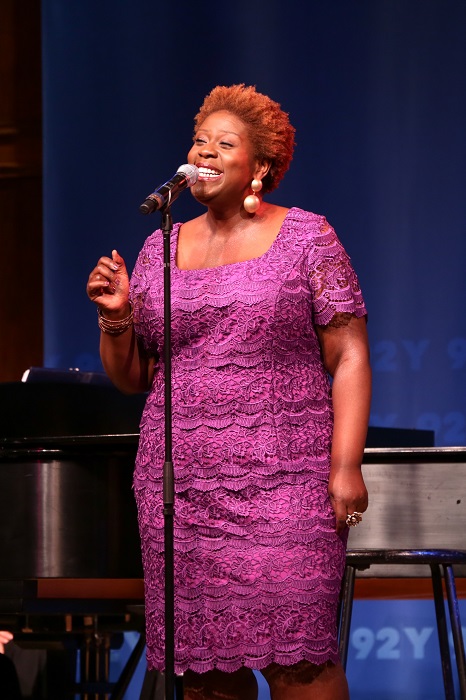 Capathia Jenkins is wearing a lavender lace dress and is singing at a microphone on a stage. Her right hand is partially raised into a fist. 