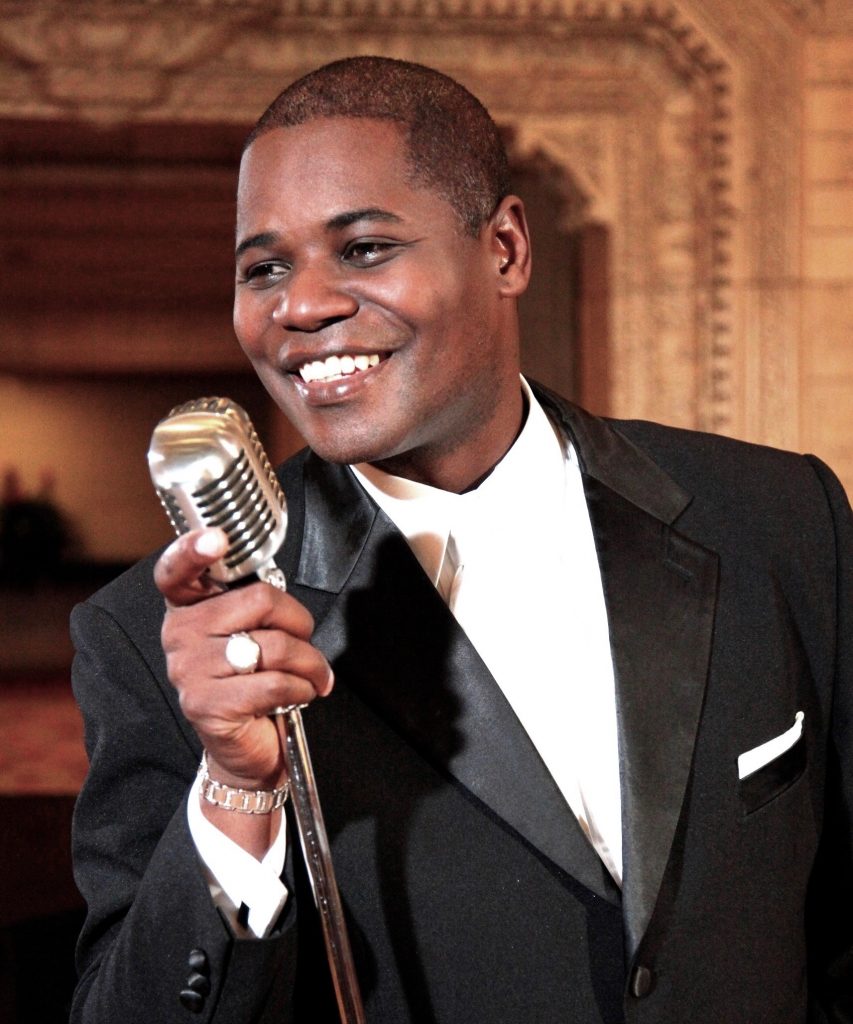 Caesar holds a retro, metal microphone and smiles brightly. He is wearing a black tuxedo jacket and white collared shirt.