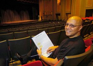 Chelsea Tipton is seated in an empty theatre holding a music score. He is looking over his left shoulder at the camera and smiling.