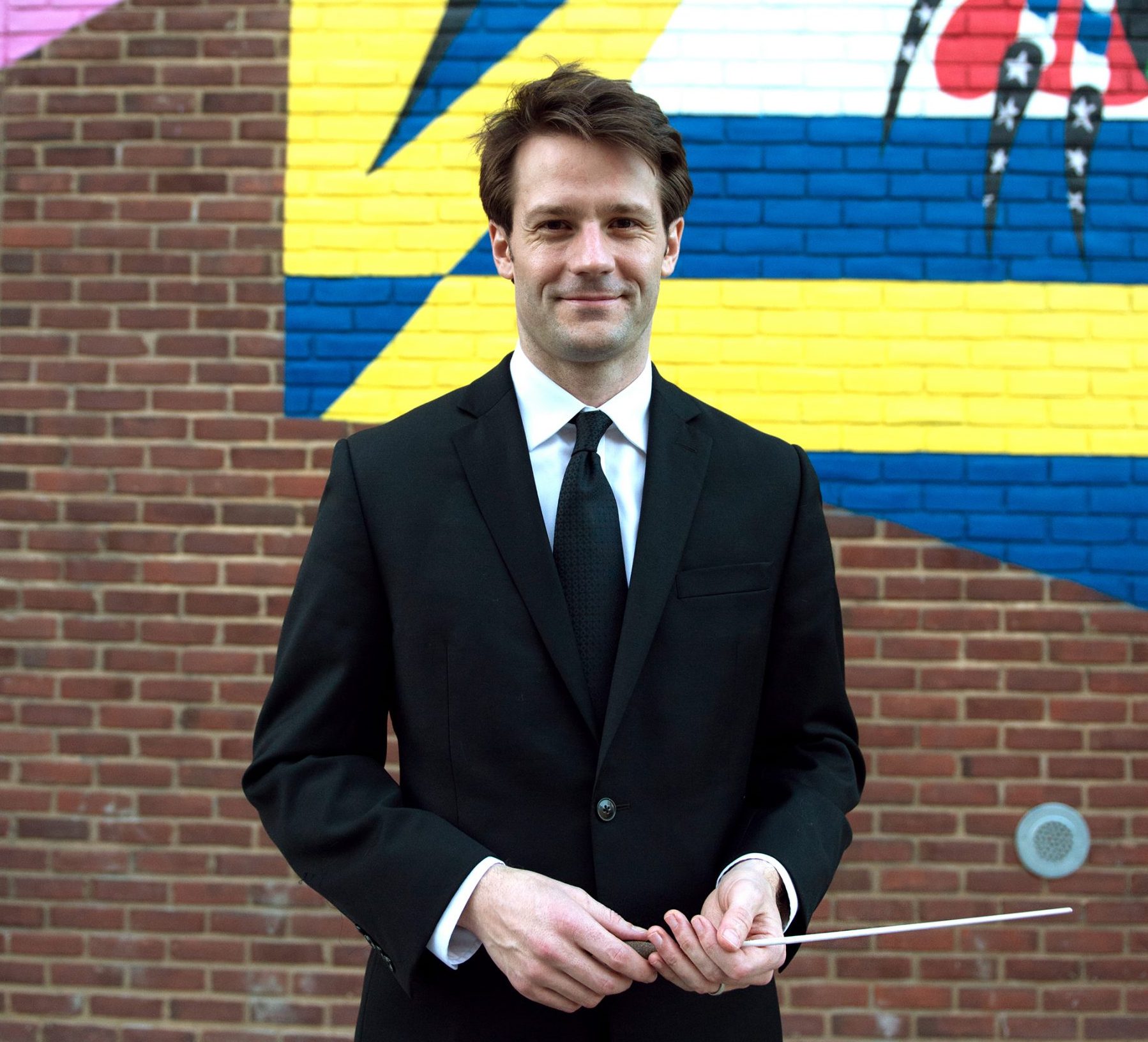 James is wearing a black suit with a white shirt and black tie. He is standing facing the camera, smiling, and is holding a conducting baton between his hands. He is in front of a multi-colored painting on a brick wall.