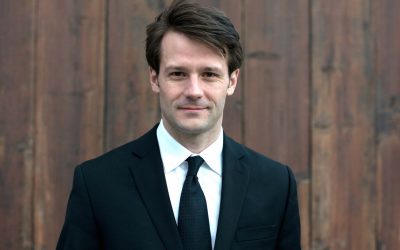 James stands in front of a wooden paneled backdrop. He is wearing a black suit and tie with a white collared shirt and his is smiling pleasantly at the camera.