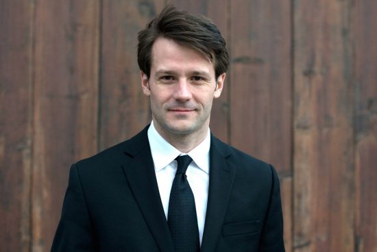 James stands in front of a wooden paneled backdrop. He is wearing a black suit and tie with a white collared shirt and his is smiling pleasantly at the camera.