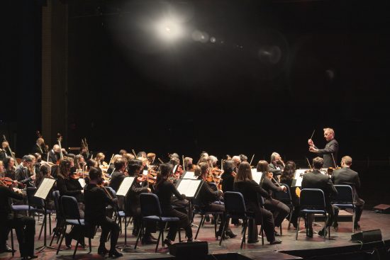 Alasdair Neale conducts the New Haven Symphony Orchestra. The orchestra is viewed from the side on the stage at SCSU. The background is completely black except for one bright spotlight shining above the orchestra.