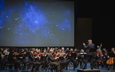 The orchestra performs on a stage in front of a large screen showing colorful lights in motion