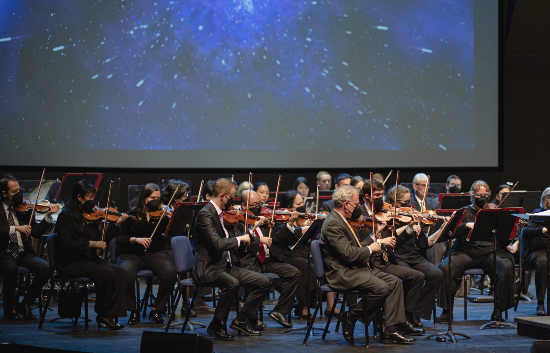 The orchestra performs on a stage in front of a large screen showing colorful lights in motion