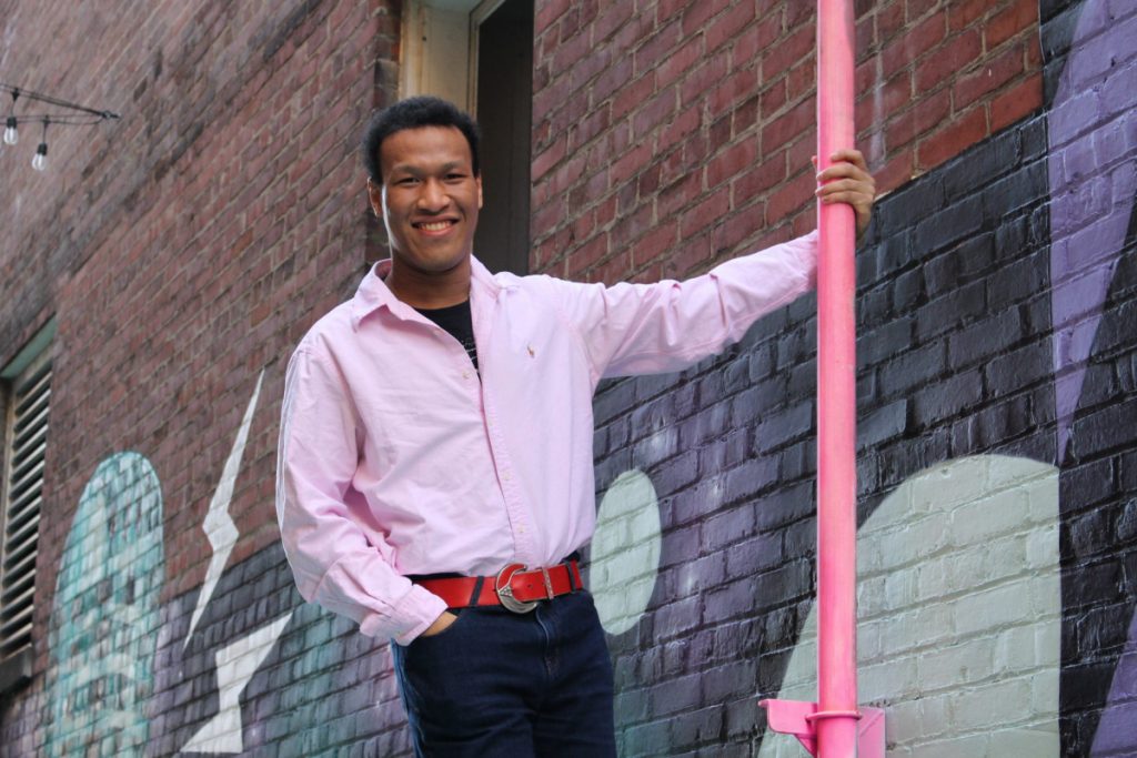 Clayton is standing in front of a brick wall. He has one hand in his pocket and the other is holding a bright pink industrial pipe. 