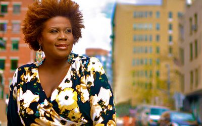 Capathia Jenkins stands in a city street. She is wearing a bright gold, white and blue top and she is looking powerfully off to her left.