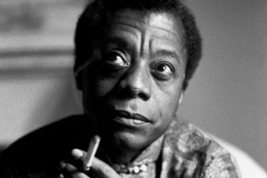 A black and white portrait of James Baldwin. Baldwin is a Black man, and in this photograph the camera is cropped close to his face. He is holding a cigarette in one hand and is glancing upwards to the right with his eyebrows raised.