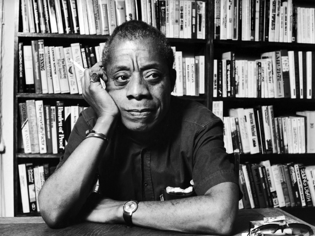 A black and white photo portrait of James Baldwin. Baldwin is a Black man and he is sitting at a table with bookshelves behind him. He has his cheek resting on his hand and his other arm across the table.