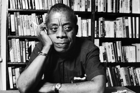 A black and white photo portrait of James Baldwin. Baldwin is a Black man and he is sitting at a table with bookshelves behind him. He has his cheek resting on his hand and his other arm across the table.