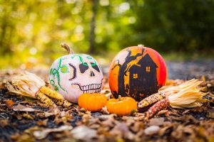 Two spooky pumpkins painted for Halloween are sitting on a pile of leaves with festive corn cobs placed on either side. There are two smaller pumpkins in the foreground of the photo, as well.
