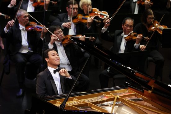 Clayton Stephenson sits a grand piano with the lid open. He is wearing a black tuxedo and has his head slightly tipped back while he plays. In the background you can see the violinists of the orchestra playing with him.