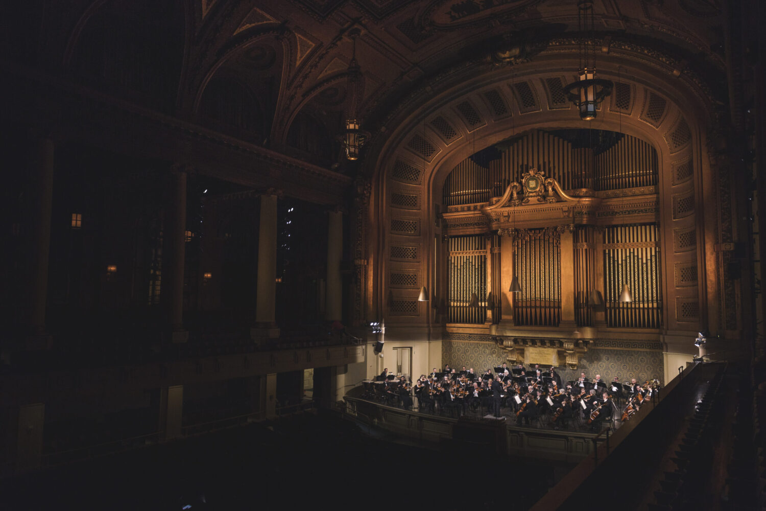 The orchestra plays on the stage at Woolsey Hall. The Hall is in shadows, and the golden organ pipes rise over the orchestra creating a moody and dynamic scene.