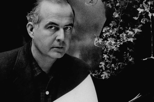 Black and white photograph of Samuel Barber posing for the camera. He is looking at something out of the image frame and has a serious expression. He is holding what are most likely music scores in his hands. He has thinning white and gray hair and pale skin.