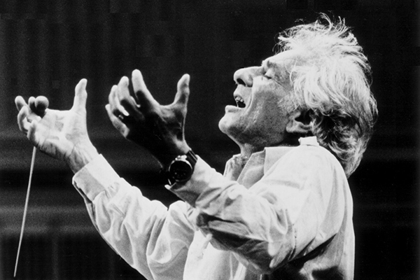 Black and white photograph of Leonard Bernstein passionately conducting an orchestra. His eyes are closed, his mouth is open, and his head is tilted backwards. In his right hand he is holding a conductor’s baton. He has shaggy, short white and gray hair and pale skin.