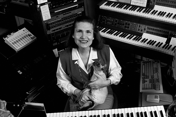 Black and white photograph of Wendy Carlos posing for the camera. She is in a recording studio surrounded by piano keyboards, synthesizers, and other instruments. She is smiling and looking upwards at the camera while a hairless cat sits in her lap. She has a shoulder-length bob and pale skin.