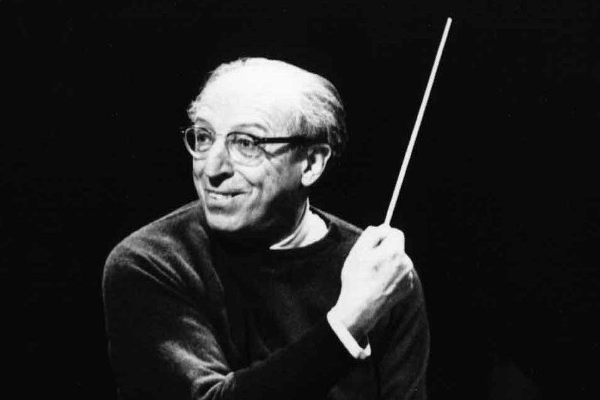 Black and white photograph of Aaron Copland conducting an orchestra. He is pursing his lips and holding a conductor’s baton in his right hand across his upper body. He has thinning white and gray hair and pale skin.