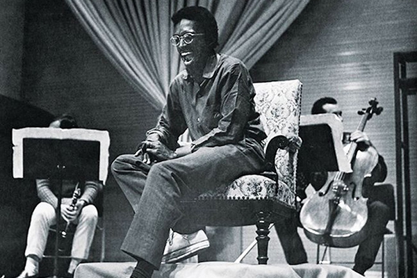 Black and white photograph of Julius Eastman. He is sitting on a chair and appears to be passionately vocalizing. His mouth is open wide and his eyes are tightly closed. He is wearing glasses with round frames and has short, thick, black hair. He has dark skin. There are two musicians sitting behind him, holding a clarinet and a cello respectively.