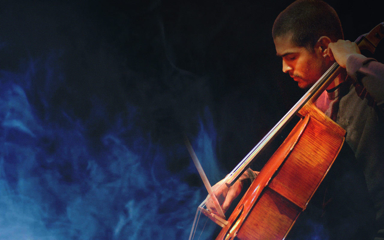 A cellist pulls his bow across his instrument, seated and shown in profile. There is a blue cloud of smoke rising from his instrument as he plays with his head down.