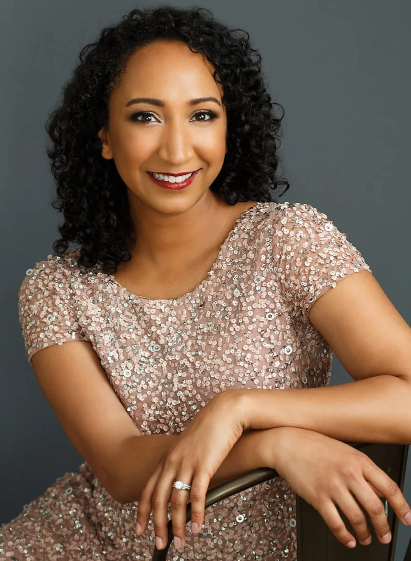 Soprano Lisa Williamson sits with her arms crossed in front of her on a chair. She is wearing a dazzling pale pink jeweled dress. 