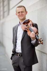David Southorn, concertmaster of the New Haven Symphony Orchestra, poses with his violin and smiles into the camera.