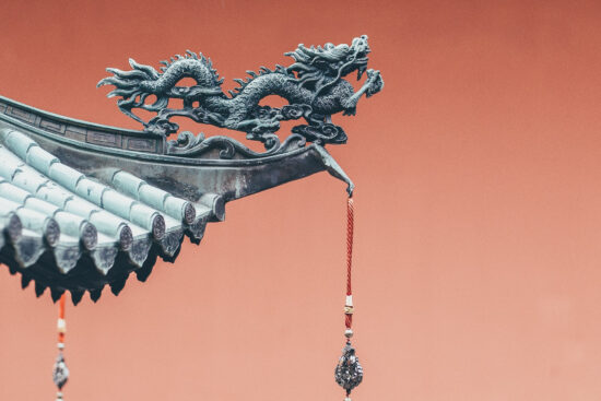 A metal statue of a dragon extends off the edge of a shingled roof. The sky is lit up a bright red/orange color.