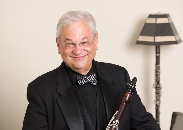 Mr. Shifrin has white, short hair and is wearing a black suit and shirt with a zebra-striped bow tie. He is holding a clarinet and behind him there is a fabulous lamp made out of a clarinet.