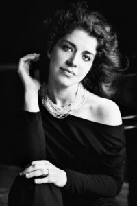 A portrait of Pianist Inna Faliks in black and white. She is shown wearing a black top with long sleeves hanging off of her shoulder and a necklace drapes in many strands around her neck. Her hair is flowing behind her and a faint outline of a piano is visible over her shoulder.