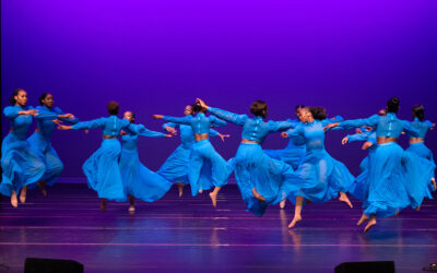 A dozen dancers in bright blue, flowing costumes, twirl across a stage under rich, purple lights.