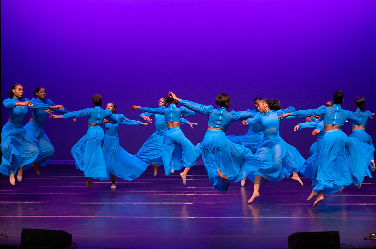 A dozen dancers in bright blue, flowing costumes, twirl across a stage under rich, purple lights.