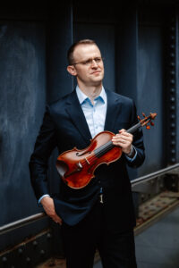 David Southorn stands holding his violin in one hand with the other in his pocket. He is wearing a dark suit with a collared shirt and looks off into the distance.