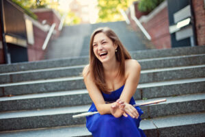 Mira sits a the bottom of a staircase wearing a bright blue dress. She has her flute on her lap and has her mouth open in a big smile.