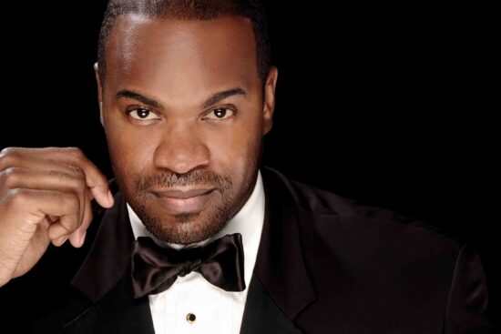 This close-up headshot shows Eric Greene, a Black man, wearing a tuxedo and adjusting his bow tie while he stares into the camera.