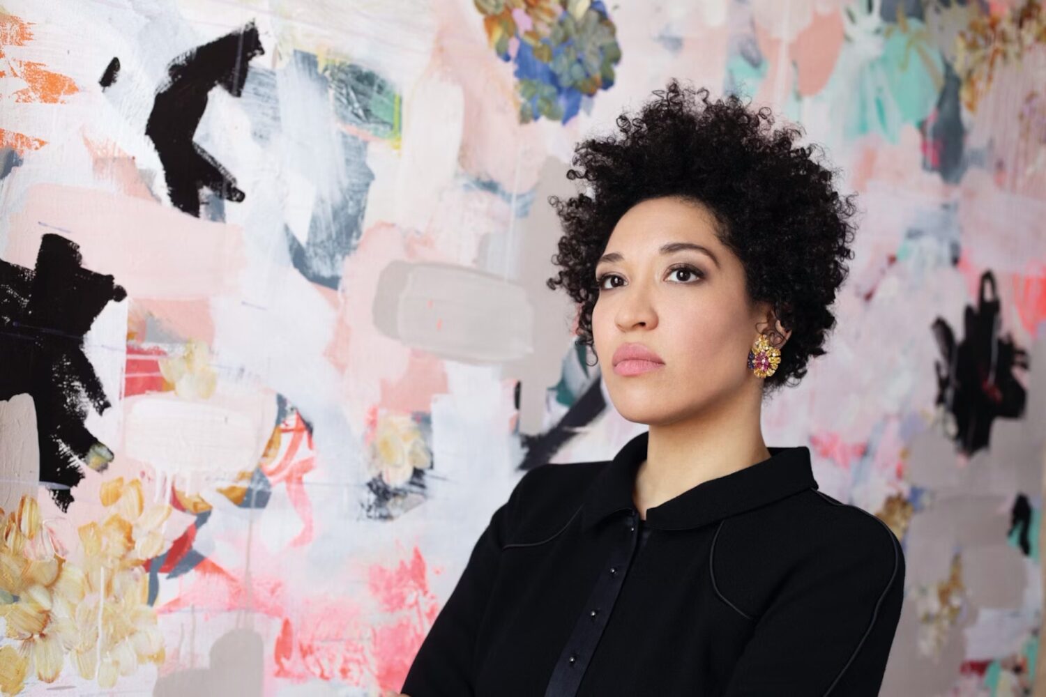 Julia Bullock, wearing a black, buttoned-up shirt, sits in front of a pastel abstract painting. She looks off into the distance.