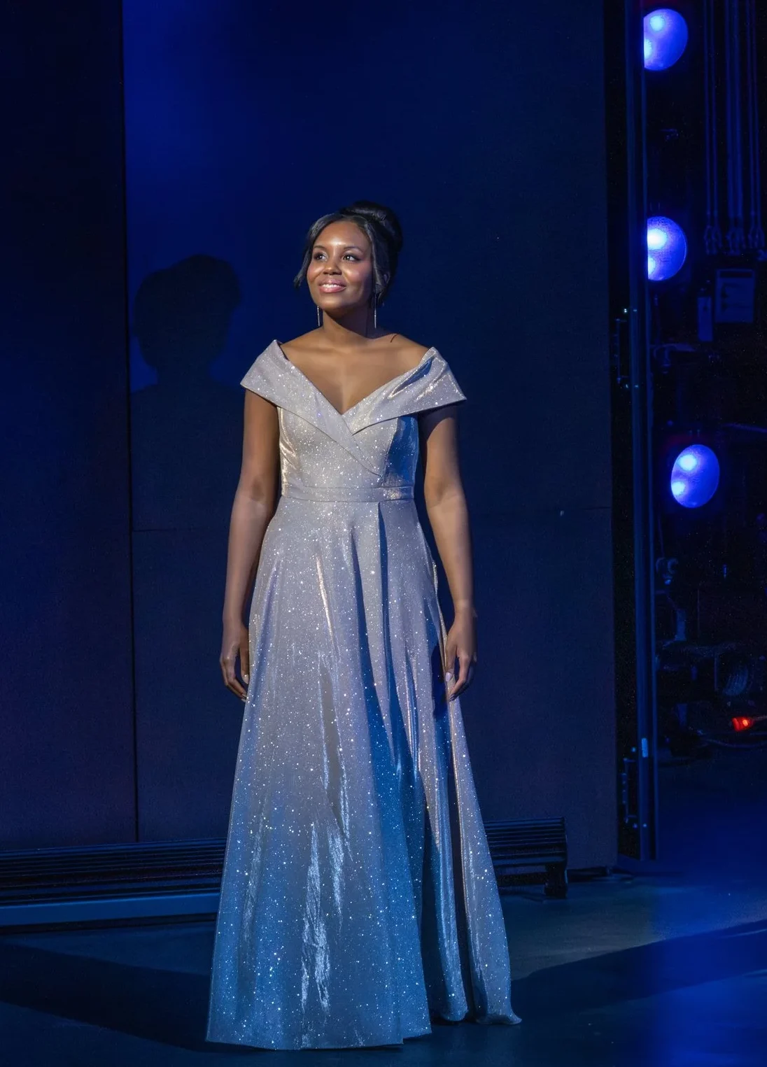Soprano Jillian Tate is wearing a sparkling gown and stands in front of blue stage lights.