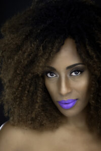 Yendi, a Black woman, is pictured up close with big brown curls and purple lipstick.