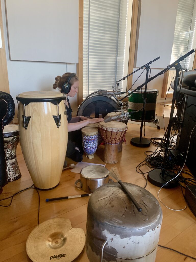 A woman with blonde hair wearing headphones sits with her eyes closed surrounded by different drums and microphones. 