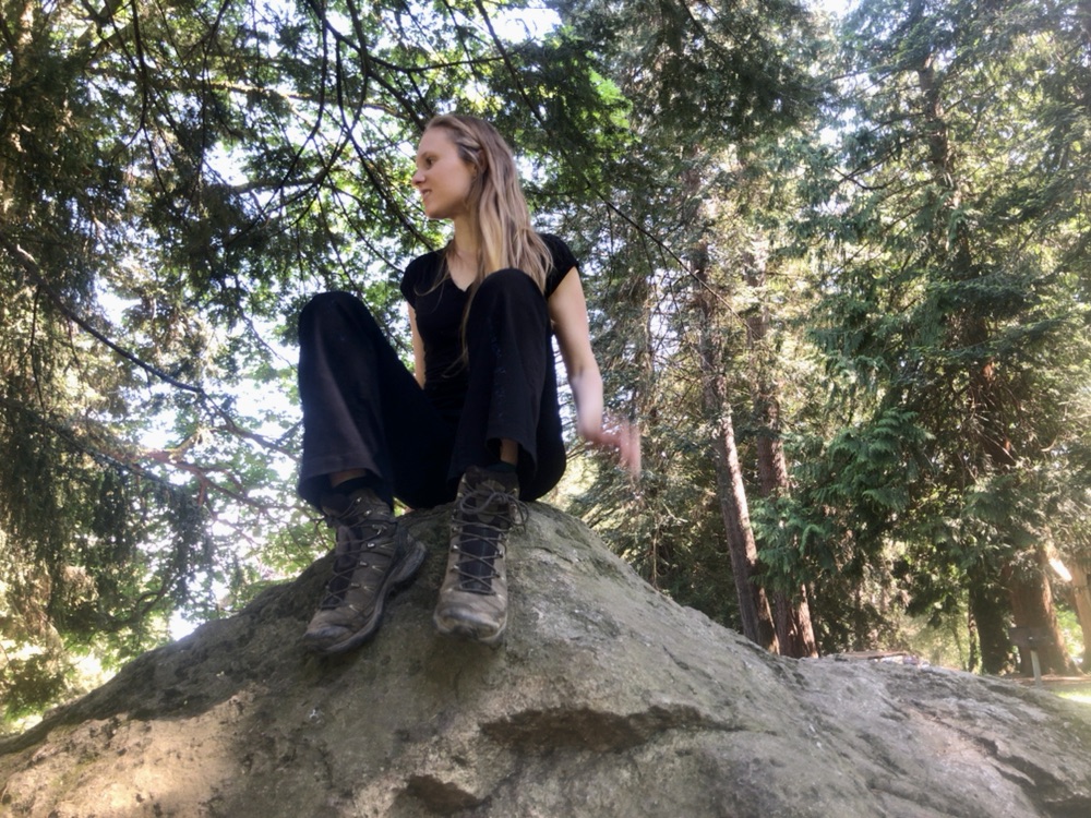 A woman with long blonde hair sits on a large rock with lots of trees in the background. 
