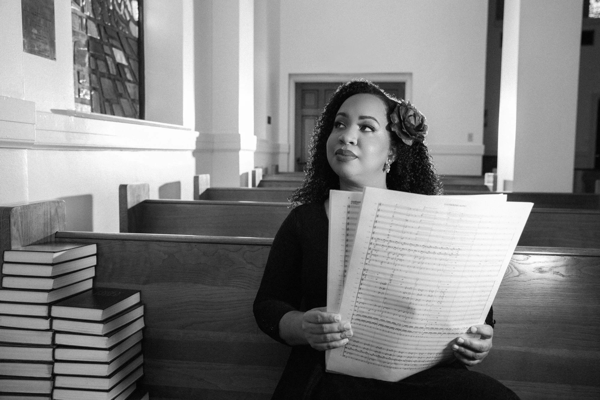 a black and white photo where a woman with shoulder length brown hair is holding sheet music looks off into the distance.