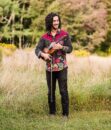 A man with shoulder length curly hair stands in a field holding a violin and looking to the side while smiling