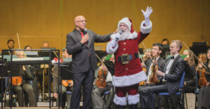 Santa Claus stands on stage in front of the orchestra and waves to the audience while holding a microphone. It is quite the holly jolly scene!