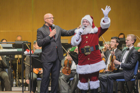 Santa Claus stands on stage in front of the orchestra and waves to the audience while holding a microphone. It is quite the holly jolly scene!